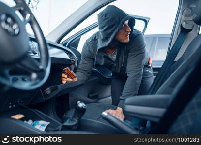 Car robber takes the wallet from the glove compartment, criminal lifestyle, stealing. Hooded male bandit opening vehicle on parking. Auto robbery. Car robber takes the wallet, stealing