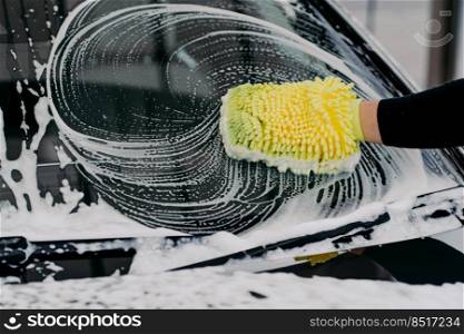 Car service concept. Unrecognizable man washes auto with cloth and car wash sh&oo, has desire to cleanliness. Hand washing vehicle with sponge