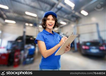 car service, job and profession concept - happy smiling female worker in blue uniform with clipboard and pen writing over auto repair shop on background. female worker with clipboard at car service