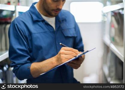 car service, repair, maintenance and people concept - auto mechanic man or smith writing to clipboard at workshop or warehouse. auto mechanic with clipboard at car workshop