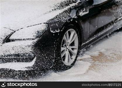 Car wash concept. Close up shot of dirty black car covered with white foam at cleaning station. Washing process. Perfect service