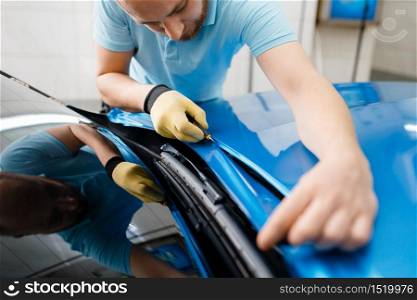 Car wrapping, man cuts protective vinyl foil or film on vehicle hood closeup. Worker makes auto detailing. Automobile paint protection coating, professional tuning. Car wrapping, man cuts protective foil or film