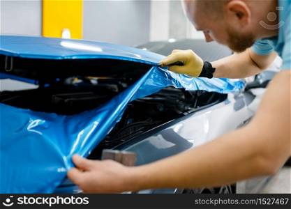 Car wrapping, man cuts protective vinyl foil or film on vehicle hood closeup. Worker makes auto detailing. Automobile paint protection coating, professional tuning. Car wrapping, man cuts protective foil or film