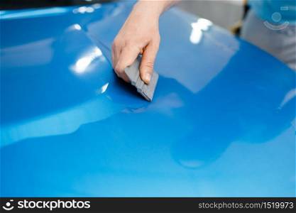 Car wrapping, mechanic with squeegee installs protective vinyl foil or film on vehicle hood. Worker makes auto detailing. Automobile paint protection coating, professional tuning. Car wrapping, mechanic with squeegee installs film