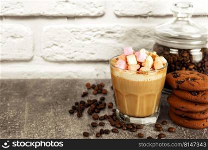 Caramel latte coffee in a glass with marshmallow top and chocolate cookie. Coffee beans on gray and white brick wall surface with copy space.. Iced caramel latte coffee in a glass with syrup and marshmallows topping