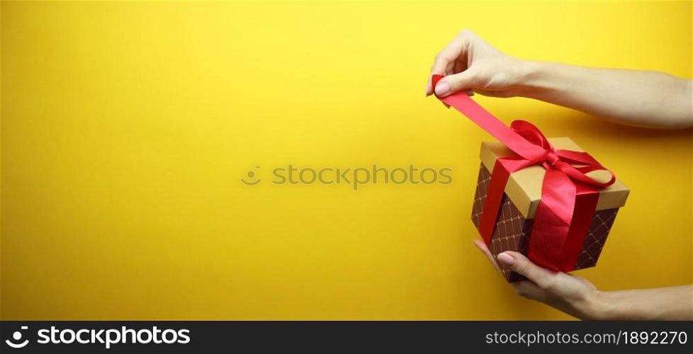 Cardboard box with a red ribbon and a bow in hands. Isolated on a yellow background. Place for your text. Copy space. Banner.
