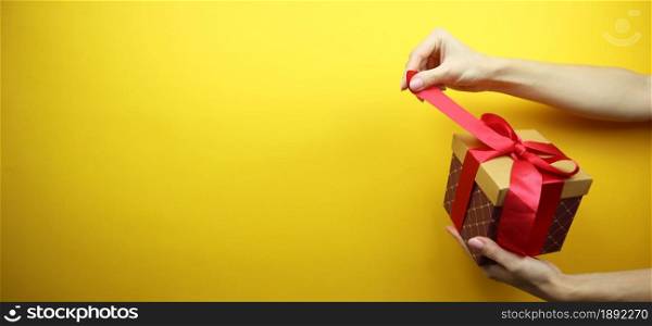 Cardboard box with a red ribbon and a bow in hands. Isolated on a yellow background. Place for your text. Copy space. Banner.