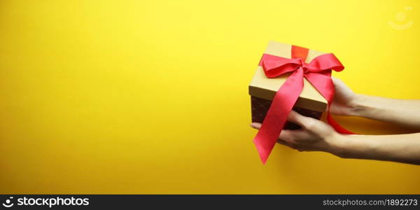 Cardboard box with a red ribbon and a bow in hands. Isolated on a yellow background. Place for your text. Copy space. Banner.. Cardboard box with a red ribbon and a bow in hands. Isolated on a yellow background. Place for your text. Banner.