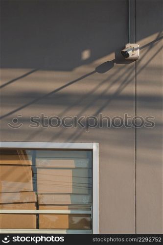 Cardboard boxes on shelf inside of glass wall frame with security camera on gray cement wall of convenience store in vertical frame, technology and security concept