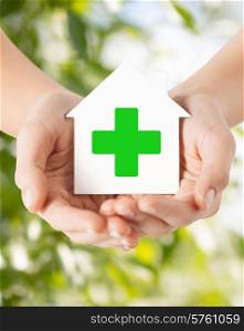 care, help, charity and people concept - close up of hands holding white paper house with green cross sign over natural background