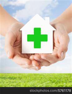 care, help, charity and people concept - close up of hands holding white paper house with green cross sign over blue sky and grass background