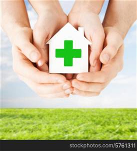 care, help, charity and people concept - close up of hands holding white paper house with green cross sign over blue sky and grass background