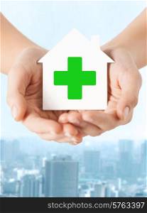 care, help, charity and people concept - close up of hands holding white paper house with green cross sign over city background