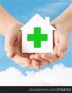 care, help, charity and people concept - close up of hands holding white paper house with green cross sign over blue sky and cloud background