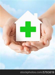 care, help, charity and people concept - close up of hands holding white paper house with green cross sign over blue sky and cloud background