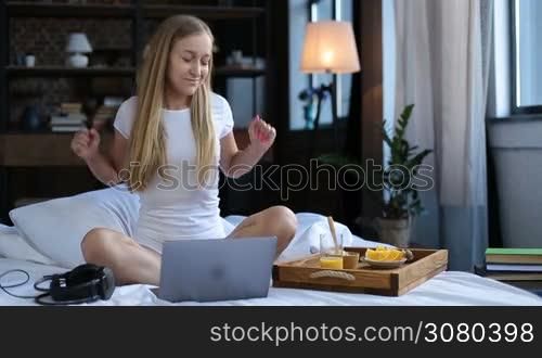 Carefree beautiful woman in white underwear sitting crossed legs on white bed sheet, listening to music with headphones during healthy morning breakfast in bed. Cheerul young female eating oatmeal, drinking orange juice and enjoying leisure at home.