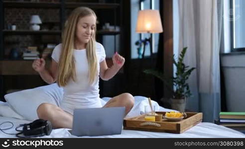 Carefree beautiful woman in white underwear sitting crossed legs on white bed sheet, listening to music with headphones during healthy morning breakfast in bed. Cheerul young female eating oatmeal, drinking orange juice and enjoying leisure at home.