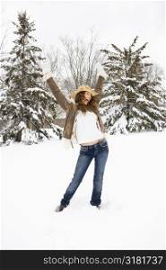 Carefree Caucasian young adult female standing with arms above head in snow wearing straw cowboy hat.