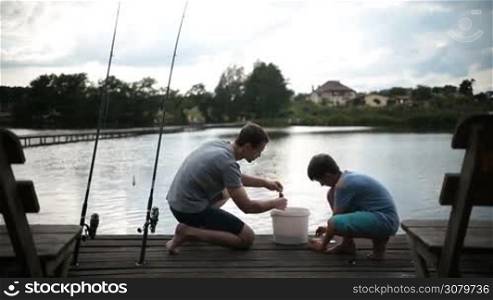 Carefree father and his teenage boy preparing to fish together, baiting fishing hook while kneeling on wooden pontoon near freshwater pond. Loving dad teaching son to bait a hook as they spend leisure angling on the lake. Back view.