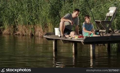 Carefree father and son sitting on wooden pontoon, enjoying meal on the lake over rural landscape background while spending weekend fishing together. Cheerful family eating breakfast during fishing at calm pond in summer. Slow motion.