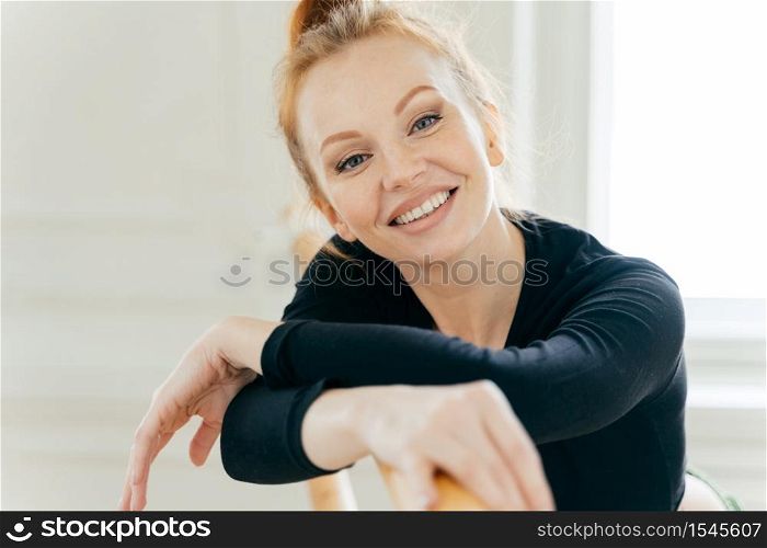 Carefree good looking ginger young female ballerina leans at ballet barre, looks gladfully at camera, practices different dancing positions, has toothy broad smile, white teeth, exercises indoor