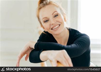 Carefree good looking ginger young female ballerina leans at ballet barre, looks gladfully at camera, practices different dancing positions, has toothy broad smile, white teeth, exercises indoor