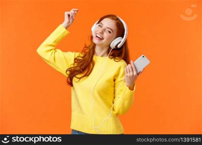 Carefree happy redhead teenage girl receive new headphones for christmas gift, dancing lift hand up while listen favorite music, holding smartphone, put awesome track, orange background.. Carefree happy redhead teenage girl receive new headphones for christmas gift, dancing lift hand up while listen favorite music, holding smartphone, put awesome track, orange background