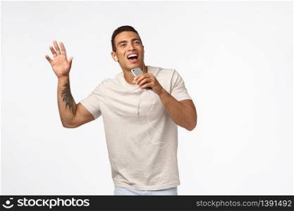 Carefree, happy smiling man in t-shirt, with tattoo, hold smartphone like microphone, singing along favorite song, raise one hand, dancing, wear wired eaprhones, play mobile karaoke, white background.. Carefree, happy smiling man in t-shirt, with tattoo, hold smartphone like microphone, singing along favorite song, raise one hand, dancing, wear wired eaprhones, play mobile karaoke, white background