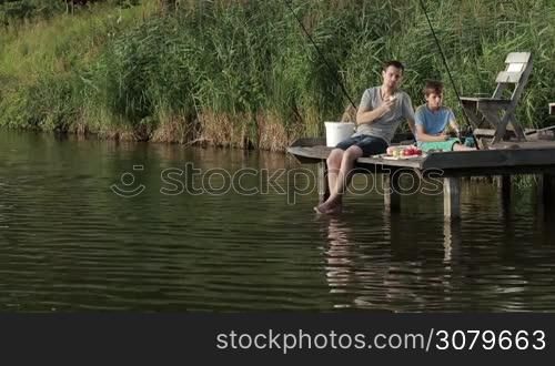 Carefree hipster father and teenage son sitting on wooden pier, enjoying meal while fishing together at calm pond against rural landscape background. Relaxed dad and boy having a snack and spending weekend fishing on the lake. Slow motion.