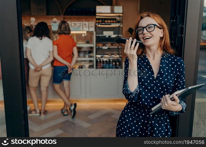 Carefree positive redhead young woman makes voice call keeps smartphone near mouth wears spectacles and polka dot dress holds laptop diary poses indoor against confectionery shop background.. Redhead young woman makes voice call keeps smartphone near mouth wears spectacles polka dot dress