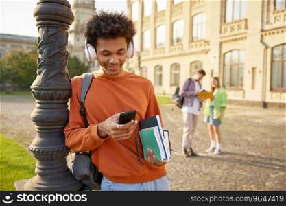 Carefree relaxed teenager boy student in earphones using mobile phone to listen music standing outdoors. Break time at high school, university or college. Carefree teenager boy student in earphones using mobile phone to listen music