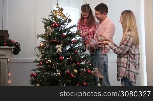 Carefree smiling daughter helping her attractive parents to decorate Christmas tree with baubles at home. Positive young father holding his cute little girl in his arms while child decorates tree during xmas holidays together with her family.