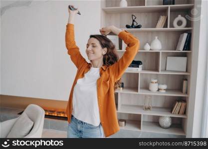 Carefree smiling female dances with closed eyes raises hands holding mobile phone listens musical playlist moving in living room enjoying sound in earphones at home. Music apps advertisement.. Girl dances holding phone listens musical playlist enjoying sound at home. Music apps advertisement
