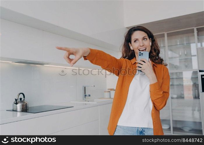 Carefree spanish woman in earphones is singing with cellphone as with microphone. Application for music listening online. Playful passionate girl is singing at home and points to you.. Carefree spanish woman in earphones is singing at home with cellphone and points to you.