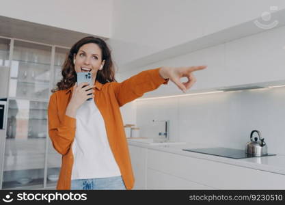 Carefree spanish woman in earphones is singing with cellphone as with microphone. Application for music listening online. Playful passionate girl is singing at home and points to you.. Carefree spanish woman in earphones is singing at home with cellphone and points to you.