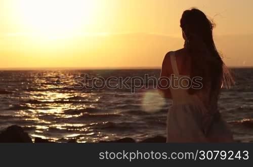 Carefree woman enjoying freedom, feeling happy and tranquil on the beach in glow of beautiful sunset. Back view. Slow motion. Serene long blonde hair female with flying scarf delighting leisure in rays of orange setting sun at seaside.