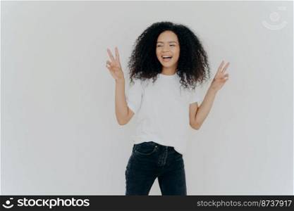 Carefree woman with Afro hairstyle raises hands up, shows victory gesture or peace sign, looks gladfully aside, dressed in casual wear, smiles relaxed, sends hello, isolated over white background.