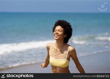 Carefree young African American female tourist, with dark curly hair in stylish swimwear, smiling happily with closed eyes while enjoying sunny summer day on sandy beach. Happy black fit woman smiling with closed eyes on sandy seashore