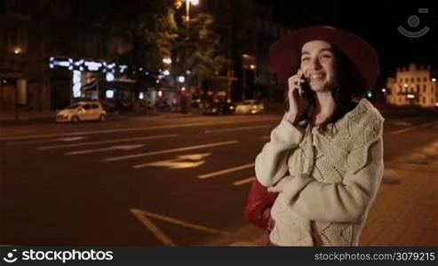 Carefree young brunette female in fashionable clothes talking on smartphone while walking along sidewalk on night city street. Gorgeous woman chatting on mobile phone while stepping down the street at night with urbanscape on background. Stabilized shot.