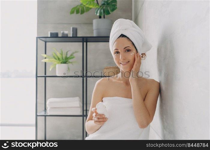 Carefree young woman is relaxing in bathroom. European lady applies face cream. Attractive caucasian girl wrapped in towel after bathing and hair washing. Happy girl at spa resort.. Carefree young woman is relaxing in bathroom. Attractive european lady applies face cream.