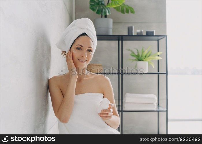 Carefree young woman is relaxing in bathroom. European lady applies face cream. Attractive caucasian girl wrapped in towel after bathing and hair washing. Happy girl at spa resort.. Carefree young woman is relaxing in bathroom. Attractive european lady applies face cream.