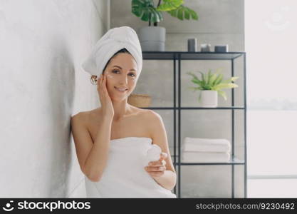 Carefree young woman is relaxing in bathroom. European lady applies face cream. Attractive caucasian girl wrapped in towel after bathing and hair washing. Happy girl at spa resort.. Carefree young woman is relaxing in bathroom. Attractive european lady applies face cream.