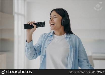 Carefree young woman wear headphones sing song to imaginary microphone, TV remote control in hand, having fun at home. Happy woman listen to music, singing, enjoying audio sound.. Excited woman in headphones sing to imaginary microphone, TV remote control, listen to music at home