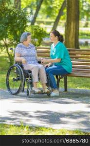 Caregiver talking to disabled senior woman in wheelchair outdoors in summer park.