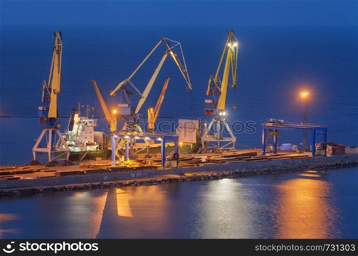 Cargo freight ship with working cranes bridge in sea port at twilight. Sea commercial port at night in Mariupol, Ukraine. Industrial view. Cargo port, logistic