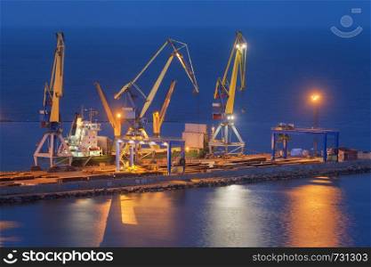 Cargo freight ship with working cranes bridge in sea port at twilight. Sea commercial port at night in Mariupol, Ukraine. Industrial view. Cargo port, logistic