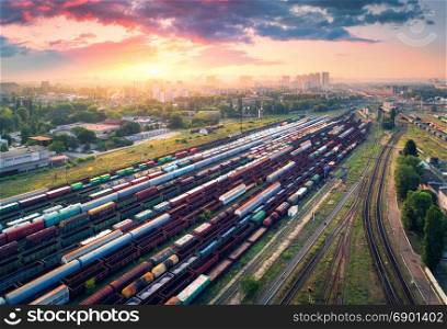 Cargo trains. Aerial view of colorful freight trains. Railway station. Wagons with goods on railroad. Heavy industry. Industrial scene with trains, city buildings and cloudy sky at sunset. Top view