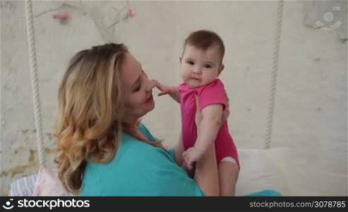 Caring charming mother kissing little fingers of her cute baby girl while enjoying time together in bedroom. Happy mom holding infant child and playing with adorable daughter while relaxing at home. Slow motion. Steadicam stabilized shot.