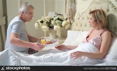 Caring elderly husband bringing breakfast to senior woman in the bedroom in the morning. Surprised wife enjoying the tasty breakfast in the bed served by her devoted man.