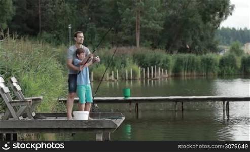 Caring father teaching son to cast fishing rod while standing together on wooden pontoon while fishing on the lake. Side view. Teenage boy throwing spinning rod from pier with the help of loving dad over beautiful rural landscape background.